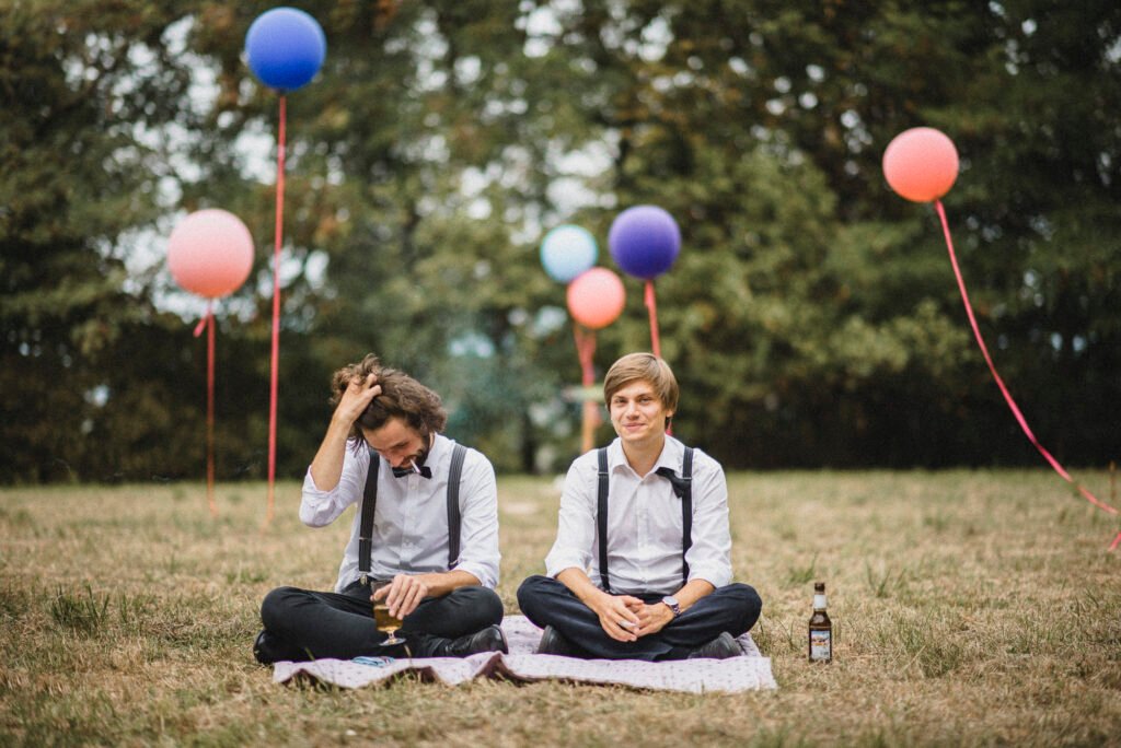 Zwei Männer mit Hosenträgern sitzen locker bei Hochzeit auf Picknickdecke im Freien